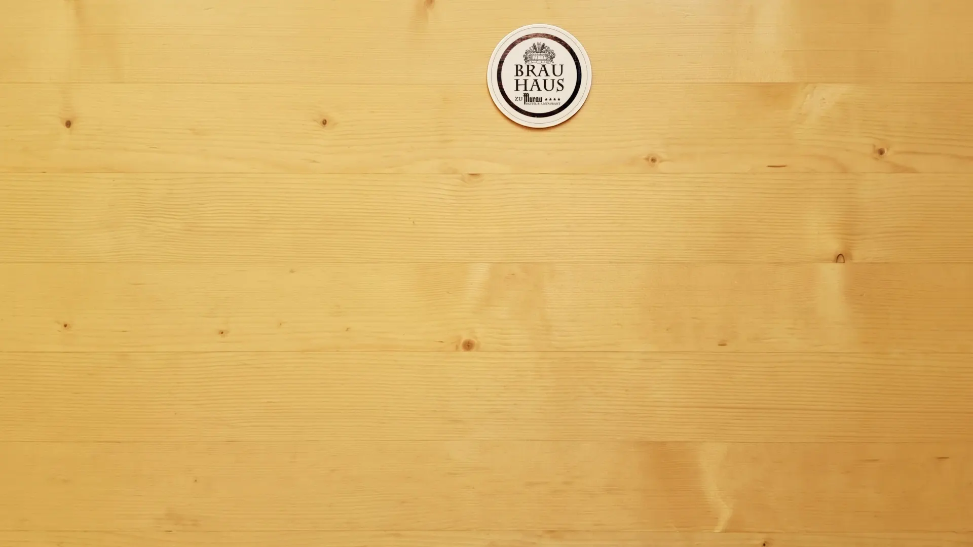 Draufsicht auf einen hellen Holztisch in der Gaststube, darauf ein Bierdeckel mit dem Logo des Brauhaus zu Murau.